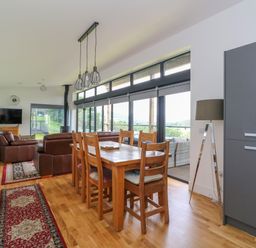 A dining table with six wooden chairs and a living area with leather sofas and a television in a room with large windows at 6 Strathtay Lodges in Aberfeldy