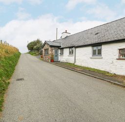 A narrow paved road with grass on the side and a white stone house with grey windows at Penrallt in Rhydlydan