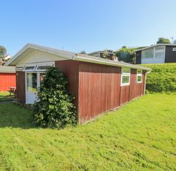 A red wooden cabin with white trim and windows surrounded by grass and nearby similar cabins at Milkwood in Elmrise Park near Llansteffan