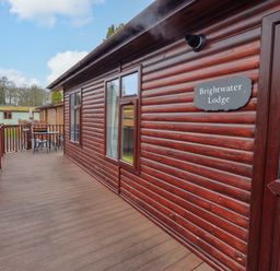 An outdoor deck with wooden railing and a table with chairs outside a wooden lodge named Brightwater Lodge at White Cross Bay near Windermere