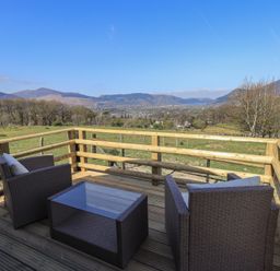 An outdoor wooden deck with two chairs and a glass top table overlooking fields and distant hills at Shepherds View in Castlerigg near Keswick