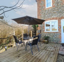 An outdoor deck with a table, four chairs, an umbrella, and a tree next to a stone and brick building at Cobble Cottage in Great Ryburgh