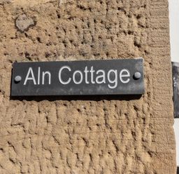 A black sign with white letters reading Aln Cottage mounted on a rough beige wall at Aln Cottage in Alnmouth