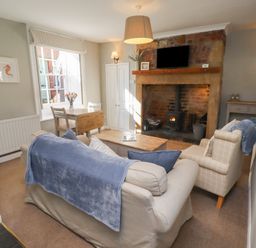 A living room with a sofa and armchair facing a fireplace with a wood stove and a wall-mounted TV at Aln Cottage (Alnmouth) in Alnmouth