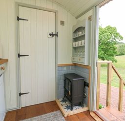 A kitchen corner with a small stove next to a door and shelves with cups and glasses at Cuckoo's Nest in South Molton