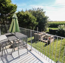 A balcony with a table and four chairs and a folded green umbrella overlooking a garden with a picnic table and barbecue at Tremornah in Lamorna
