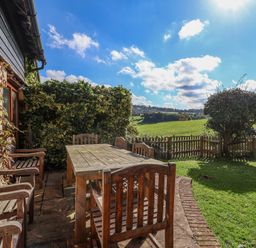 An outdoor patio with wooden table and chairs overlooking a grassy garden and field at Highgrove Barn in Painswick