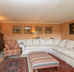 A living room with a large white sectional sofa a patterned ottoman a plaid armchair framed pictures on the wall and a fireplace at Highgrove Barn in Painswick