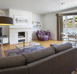 A living room with a gray sofa, glass coffee table, fireplace, wall art, TV, purple armchair, dining table, and large windows overlooking a waterfront at 21 Dart Marina in Dartmouth