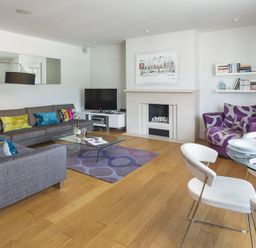 A living room with two gray sofas colorful pillows a glass coffee table a purple armchair a glass dining table with white chairs and a fireplace at 21 Dart Marina Dartmouth