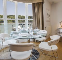 A dining area with a round glass table set with plates and glasses near large windows showing a marina at 21 Dart Marina in Dartmouth