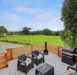 An outdoor patio with black wicker chairs a small table a chiminea a barbecue grill and a wooden fence overlooking green fields at The View in Bangor-On-Dee
