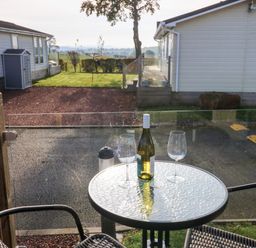 A patio table with a bottle and two glasses between two chairs with views of a driveway and houses at Brocklebank Lodge 35 High Hesket near Armathwaite