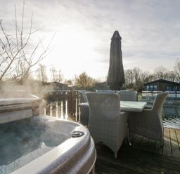 An outdoor seating area with a table and four wicker chairs next to a steaming hot tub on a wooden deck by the water at The Three Bears Lodge Coniston Drive 11 South Lakeland Leisure Village near Warton