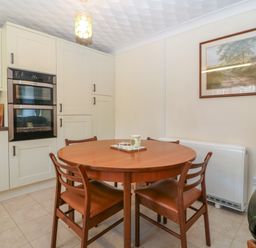 A kitchen with a round wooden dining table and four chairs a double oven and wall cabinets with a landscape painting on the wall at Gwynlys in Cemaes Bay