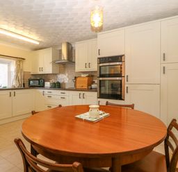 A kitchen with a round wooden table and chairs with white cabinets and built in oven at Gwynlys in Cemaes Bay