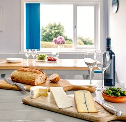 A kitchen countertop with bread cheese olives wine and wine glasses with a dining table and window in the background at Carousel in Broad Haven