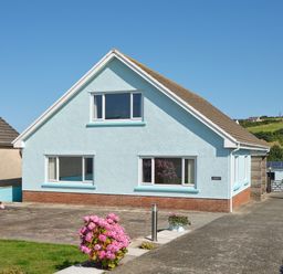 A light blue house with a sloped roof and large windows with pink flowers in front at Carousel in Broad Haven
