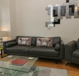 A living room with two gray sofas with sheep pillows a glass coffee table with books a lamp on a side table and a geometric mirror on the wall at 12 Blue Hill Park in Ambleside
