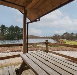 A wooden picnic table and bench on a covered porch overlooking a lake with trees on the shore at Kingfisher Lodge in Treverbyn near St Austell