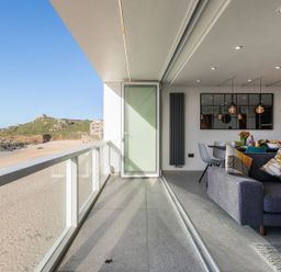A balcony overlooking a sandy beach and rocky hill with a living room and dining area visible through open glass doors at Louies Loft in St Ives