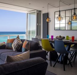 A living room with a gray sofa and colorful pillows next to a dining table with multicolored chairs and ocean view at Louies Loft in St Ives