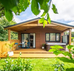 A wooden cabin with a small porch table and chairs a hot tub and greenery around at Maple Lodge in Angarrack
