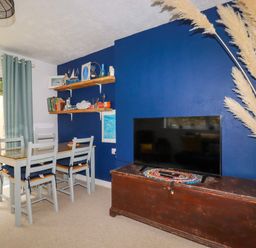 A dining table with four chairs near sliding glass door and TV on wooden chest against blue wall at 1 Mill Cottages in Porthtowan