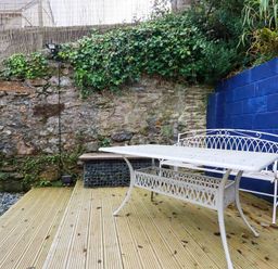 An outdoor seating area with a white metal table and bench on a wooden deck next to stone and blue painted walls with greenery at 1 Mill Cottages in Porthtowan