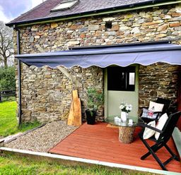An outdoor patio area with a stone wall house a green door a glass table on a wooden base two black chairs with cushions and a retractable awning at The Old Workshop in Dre-Fach near Lampeter
