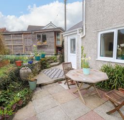 A small garden patio with a wooden table and two chairs next to a house and a wooden shed in the background at Elder Cottage Studio in Perranwell Station near Ponsanooth
