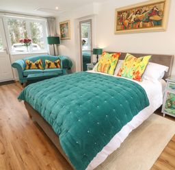 A bedroom with a bed a teal comforter colorful pillows a teal sofa lamps and paintings at Elder Cottage Studio in Perranwell Station near Ponsanooth