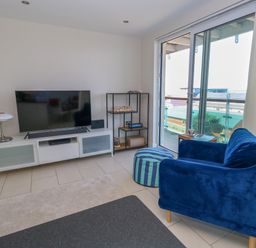 A living room with a blue armchair a striped pouf white tv stand with a television and a glass door leading to a balcony at Manhattan Beach in Castletown Portland