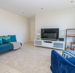A living room with a blue sofa, television on a white stand, and a metal shelf with decorative items at Manhattan Beach in Castletown Portland