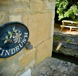 A stone wall with a decorative sign saying Windrush and a small bell next to a stone ledge and a wooden bench under trees at Windrush in Burford