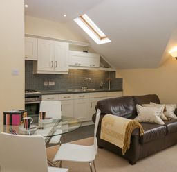 A kitchen and dining area with a glass table and white chairs and a brown leather sofa with cushions near a floor lamp at Ramblers Lodge Bowland Bridge near Bowness-On-Windermere Lake District