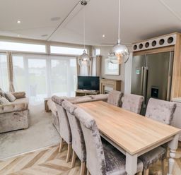 A dining area with a wooden table and six chairs next to a kitchen with a fridge and kettle and a living area with sofas and a TV at Finch 65 in Cayton Bay