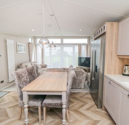 A dining table with chairs next to a kitchen area with a refrigerator and a kettle in a living space with sofas and a TV at Finch 65 in Cayton Bay