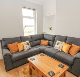 A living room with a grey corner sofa with orange and bee-themed cushions a wooden coffee table and a window at Grace House in Silloth