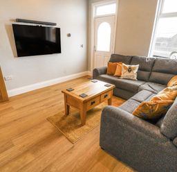 A living room with a grey sectional sofa wooden coffee table mounted flat screen tv and wooden door at Grace House in Silloth