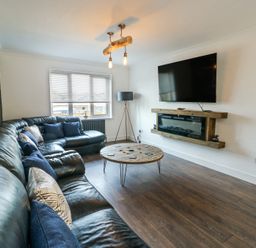 A living room with a black leather corner sofa a round wooden coffee table a wall-mounted TV and an electric fireplace at Ty Copr in Amlwch