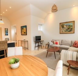 An open living room and kitchen area with a round wooden table and chairs a corner sofa with cushions and a small wooden coffee table a TV on a stand and wall paintings at The Elm Coach House in Chipping Norton