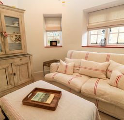 A sitting room with sofa, cushions, wooden cabinet, and ottoman with tray and book at The Elm Coach House in Chipping Norton