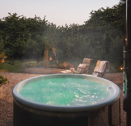An outdoor hot tub with bubbling water on a gravel patio next to two lounge chairs and greenery at Moonshine in St Erme near Truro