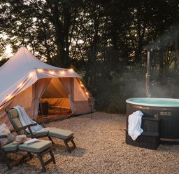 A tent with string lights beside two padded lounge chairs and a hot tub in a wooded outdoor area at Moonshine in St Erme near Truro