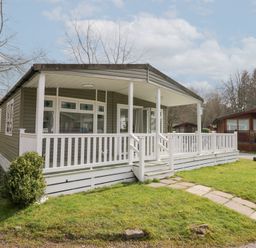 A cabin with a covered porch and white railing in a grassy area at Cosy Corner in White Cross Bay near Troutbeck Bridge
