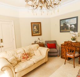 A sitting room with a cream sofa and floral cushions a gray armchair with a red cushion a wooden desk with a chair and lamp and two paintings on the walls at Hereford House in Tenby