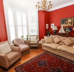 A living room with red walls, a beige sofa, striped chairs, a wicker chair, a patterned rug, lamps, and framed artwork at Hereford House in Tenby