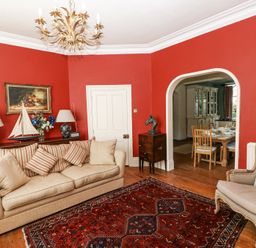 A living room with beige sofa and armchair red walls wooden floor and an archway to a dining room at Hereford House in Tenby