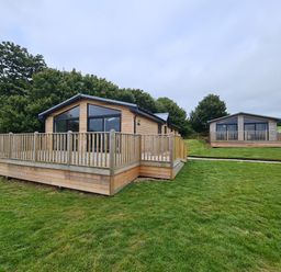 Two wooden cabins with large windows and raised decks on a grassy area with trees at Bay in Ilfracombe